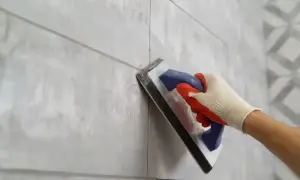 Ręka w rękawicy nakłada fugę na szare płytki za pomocą kielni.