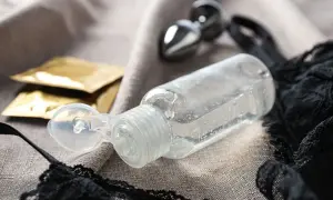 Butelka lubrykantu, dwa opakowania prezerwatyw i metalowy dildo na tle czarnej koronki i szarej tkaniny.