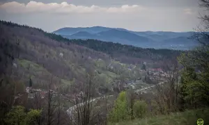 Widok na dolinę z zabudowaniami, lasami i pasmem górskim w tle. Widać tu ćwilin z Jurkowa, wiosenną zieleń i chmury na niebie.