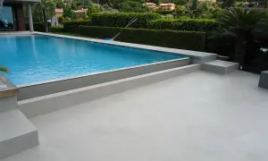 Modern villa with a bright blue pool and surrounding terraces finished with elegant piscinas de microcemento. Lush green hedges and palm trees frame the scene.
