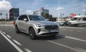 Srebrny Volvo XC90 T8 w ruchu miejskim, z nowoczesnymi budynkami w tle.