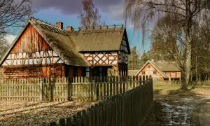 Drewniane domy z krytymi strzechą dachami i drewnianym płotem na tle drzew.