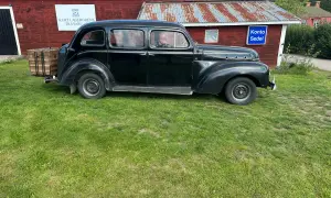 Svart Volvo PV 831 parkerad på gräs framför en röd byggnad med rostigt plåttak. En träkorg sitter på bakre stötfångaren.