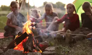 Kinder rösten Marshmallows über einem Lagerfeuer. Ein Zelt ist im Hintergrund zu sehen.