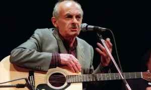 Starszy mężczyzna z gitarą i mikrofonem, śpiewający piosenkę o wojnie.
