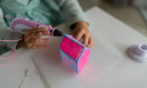 Dziecko tworzy różowy domek z niebieskim dachem za pomocą długopisu 3D. Powstaje piękny, trójwymiarowy ornament.
