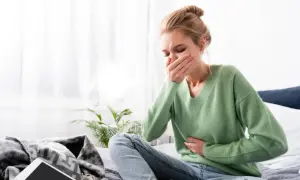 Mujer rubia con moño sentada en la cama, con la mano en la boca y otra en el abdomen, sufriendo nauseas antes de la regla.