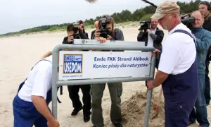 Na plaży nudystów stogi piasku i tablica informacyjna. Ludzie z aparatami dokumentują wydarzenie.