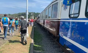 Gente con chalecos amarillos espera el tren descenso del Sella. El tren tiene vagones marrones y azules con detalles dorados.