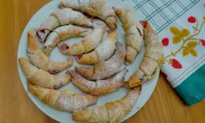 Kruche rogaliki z marmoladą, posypane cukrem pudrem, ułożone na talerzu.