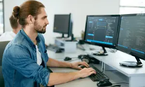 Młodszy programista z kitką na głowie pracuje przy dwóch monitorach wyświetlających kod.