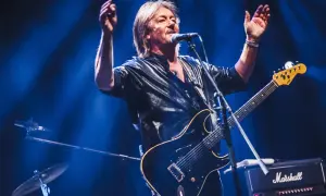 Chris Norman, lider zespołu Smokie, śpiewa i gra na gitarze na scenie, oświetlony niebieskim światłem.
