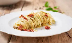 Un piatto di linguine condite con pomodori secchi, capperi e prezzemolo, su un piatto bianco con sfondo in legno.