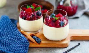 Zwei Gläser cremiger Panna Cotta mit Beerenkompott, ein köstliches rezept panna cotta original italienisch. Blaue Serviette und Holzlöffel mit Blaubeeren.