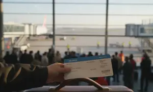 Ręka trzyma kartę pokładową z napisem "PRIORITY" na tle lotniska z samolotem i pasażerami.