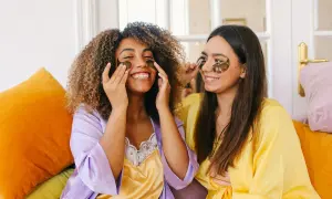 Due amiche sorridono mentre applicano patch per gli occhi, un essenziale skincare step, indossando vestaglie colorate.