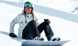 Uśmiechnięta snowboardzistka w kasku i goglach siedzi na śniegu obok deski. Jej pokrowiec na deskę snowboardową jest gotowy do drogi.