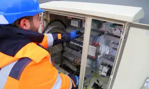 Elektryk w kasku i pomarańczowej kurtce pracuje przy skrzynce z przewodami. To jego zadanie, by wszystko działało sprawnie, jak w sep g1.