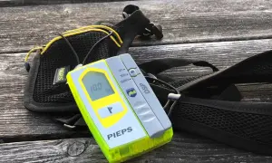 Ein Pieps lawinen sender mit Display und Suchfunktion, daneben eine schwarze Tasche mit gelben Akzenten, liegt auf einer Holzoberfläche.