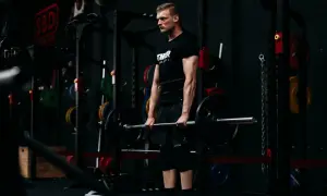 Mann mit Bart hebt eine Langhantel im Fitnessstudio. Er trägt ein schwarzes T-Shirt und kurze Hosen, ein Gürtel stützt seinen Rücken. Ein Griffkraft Trainer, der seine Leistung steigert.