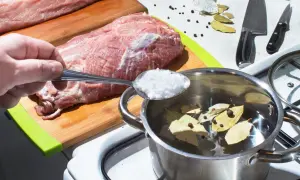 Przygotowanie solanki do mięsa: ręka wsypuje sól do garnka z wodą, liśćmi laurowymi i pieprzem. Obok surowe mięso na desce.