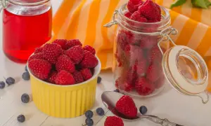 Sok z malin przepis babci: świeże maliny w żółtej miseczce i słoiku, obok czerwony napój i borówki.