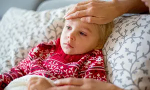 Małe dziecko w czerwonym sweterku z wzorem płatków śniegu leży chore, z gorączką. Ręka opiekuna sprawdza temperaturę. To typowy obrazek, gdy dopada nas wirus.