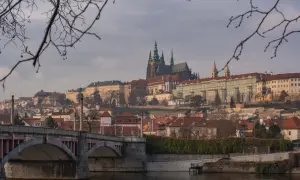 Praga nietypowe atrakcje: widok na Hradczany i Most Karola przez Wełtawę, z gałęziami drzew w pierwszym planie.