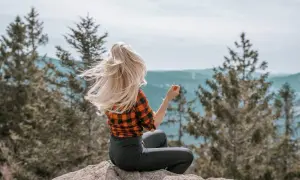 Frau mit wehendem blonden Haar sitzt auf einem Felsen im bayerischen Wald und genießt die Aussicht.