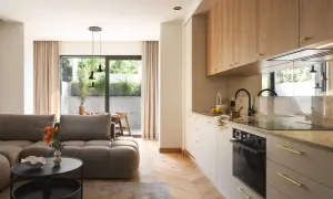 Nowoczesny salon z aneksem kuchennym, szara sofa, drewniane meble, okno z widokiem na ogród.