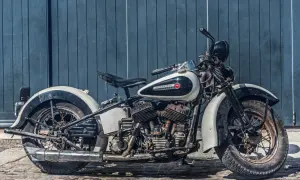 Stary motocykl Harley-Davidson w kolorze białym i czarnym, stojący na bruku przed niebieską, drewnianą ścianą.