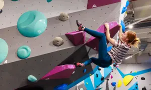 Frau klettert an einer bunten Wand in einer Boulderhalle in Hamburg. Sie hält sich an einem pinken Griff fest.