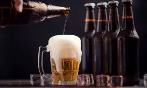 Una mano versa birra in un bicchiere traboccante di schiuma. Sullo sfondo, bottiglie di ibu birra e cubetti di ghiaccio.