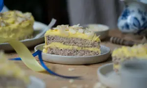 Ein Stück schwedische Mandeltorte mit gelber Creme und Mandelblättchen, dekoriert mit blauen und gelben Bändern.