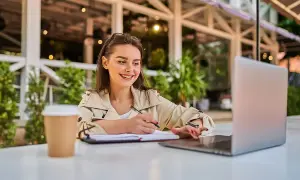 Uśmiechnięta młoda kobieta pracuje przy laptopie i notuje. Idealny przykład zatrudnienia młodocianego na umowę zlecenie.