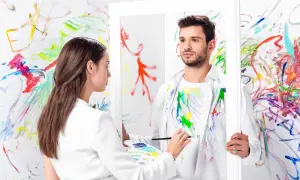 Kobieta maluje mężczyznę w lustrze, tworząc dzieło sztuki nowoczesnej. Tło jest pokryte kolorowymi pociągnięciami pędzla.