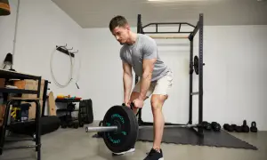 Homme musclé en buste penché, soulevant une barre chargée de poids dans une salle de sport à domicile.