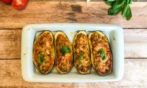 Quatre moitiés d'aubergines farcies, gratinées et garnies de basilic, dans un plat blanc sur fond de bois.