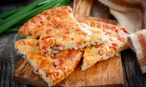 Pyszna tarta z kiełbasą, pokrojona na kawałki, leży na drewnianej desce. Obok zielona cebulka i ręcznik.