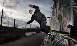 Martin Ander gör ett trick på skateboard i en betongpark med graffitiväggar och järnvägsspår i bakgrunden.