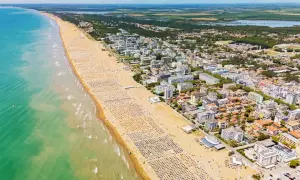 Słoneczna bibione plaża z tysiącami leżaków i parasoli, turkusowe morze i miasto w tle.