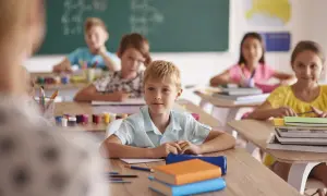 Dzieci siedzą w klasie przy ławkach, przed nimi książki i przybory.