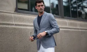 Ein Mann mit einem grauen Sakko kombiniert es lässig mit einem dunklen T-Shirt und weißen Hosen. Er hält eine Sonnenbrille in der Hand.