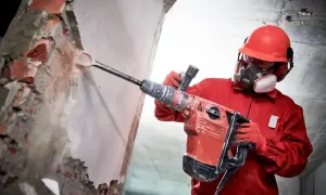 Robotnik w czerwonym kombinezonie i masce ochronnej używa maszyny do skuwania tynku, rozbijając ścianę z cegieł.