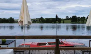 Gemütliches Restaurant am See mit rotem Tretboot und Sonnenschirmen. Die Uferlandschaft ist grün und hügelig.