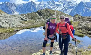 Dwie kobiety cieszą się trekkingiem wokół Mont Blanc. Mają plecaki i kije trekkingowe, a w tle widać ośnieżone szczyty.