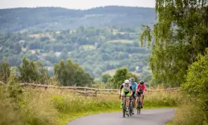 Trzech rowerzystów jedzie po asfaltowej drodze wśród zielonych wzgórz.