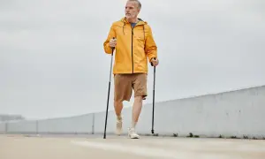 Mężczyzna w żółtej kurtce i brązowych spodenkach wykonuje rozgrzewkę przed nordic walking, trzymając kije.