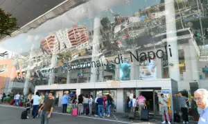 Ludzie z bagażami przed nowoczesnym budynkiem Aeroporto Internazionale di Napoli.