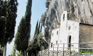 Piccolo eremo di San Valentino incastonato nella roccia, circondato da alti cipressi e un cielo azzurro.